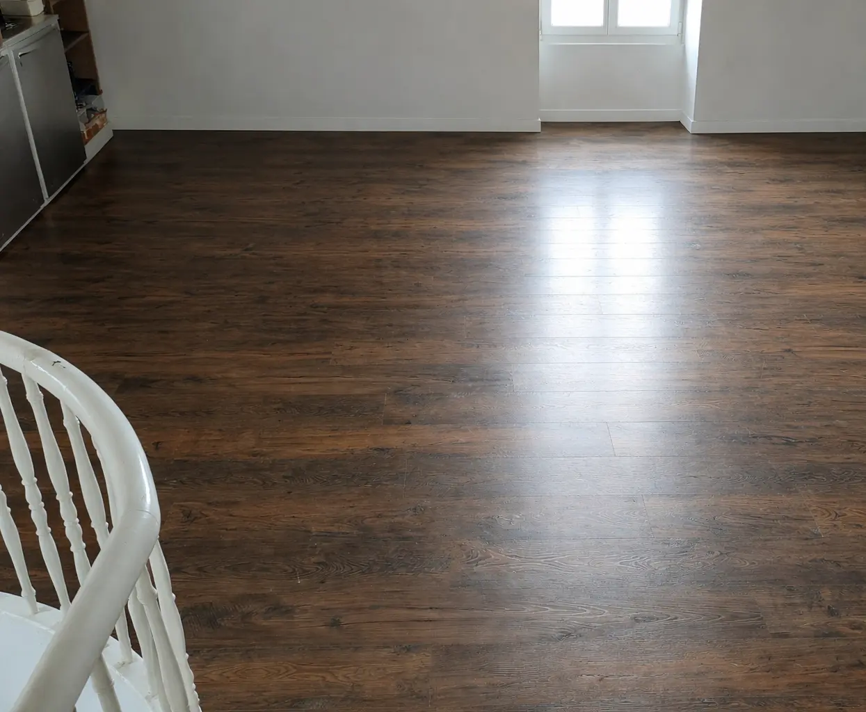 Grand espace rénové à l'étage d'une maison à Saint-Jean-de-Luz, doté d'un sol en PVC imitation bois foncé à l'aspect texturé, à côté d'un garde-corps d'escalier blanc, sous la lumière naturelle d'une fenêtre.