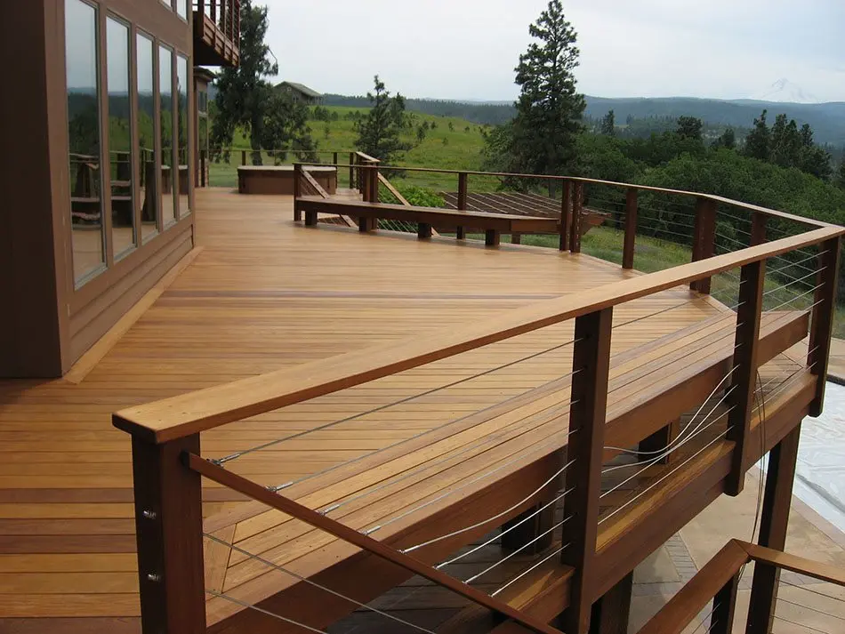 Aménagement extérieur d'une villa moderne à Urrugne avec une vaste terrasse en lames de bois exotique, aménagée avec un salon de jardin en rotin, des jardinières contemporaines et des garde-corps en verre, offrant une vue panoramique sur les montagnes de la Rhune en arrière-plan.
